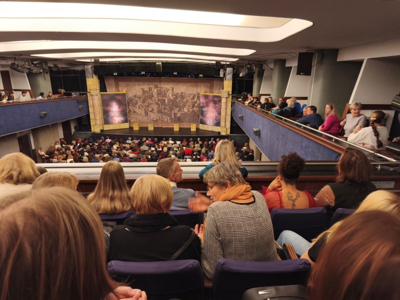 Fotografie zachycuje divadelní sál plný diváků čekajících na začátek představení, při pohledu z balkonu na oponu s motivem starověkého města.